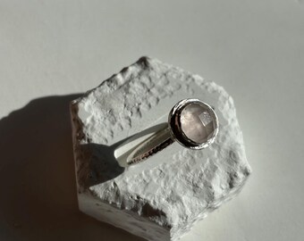Puede incluir: Un anillo de plata con una piedra preciosa rosa pálida. El anillo tiene una banda texturizada y un engaste de bisel.
