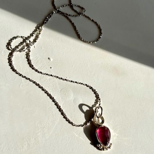 May include: A silver chain necklace with a pink gemstone pendant. The pendant is set in a silver bezel and has a delicate design.