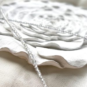 Puede incluir: Un delicado collar de cuentas blancas con cuentas iridiscentes, sobre un fondo de tela blanca.