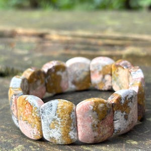 Puede incluir: Una pulsera elástica compuesta por cuentas de piedra rectangulares y lisas con un patrón moteado en tonos rosa suave, marrón tierra, crema y gris, que presenta intrincadas inclusiones fósiles en forma de estrella en toda la superficie pulida.