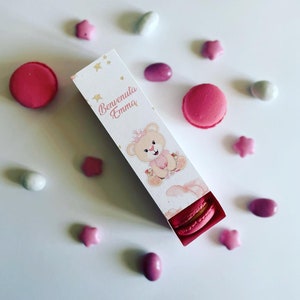 Op de afbeelding: Een witte doos met een roze en gouden teddybeerontwerp. De doos heeft de tekst "Benvenuta Emma" en bevat roze macarons. De doos is omringd door roze en witte snoepjes.