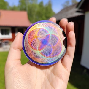Puede incluir: Una pegatina redonda e iridiscente con un diseño geométrico colorido. La pegatina tiene un esquema de color azul, morado, naranja y amarillo. El diseño es intrincado y detallado, con una variedad de líneas y formas.