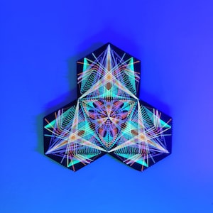 Puede incluir: Arte mural geométrico compuesto por tres paneles hexagonales negros. Cada panel presenta un diseño complejo de arte de cuerdas colorido en tonos de azul, verde, naranja y morado, creando un patrón simétrico vibrante. El fondo es un degradado de azul.