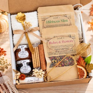 May include: A gift box containing two pouches of loose-leaf tea labeled Hibiscus Mint and Relax & Restore, two small jars of honey and raspberry jam, a gold flower-shaped spoon, a gold tea infuser, cinnamon sticks, and a Warm Vanilla lip balm.