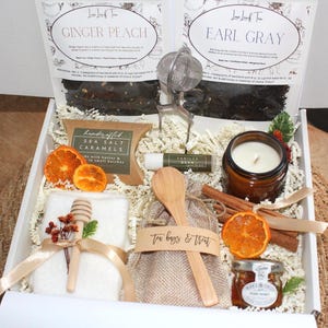 May include: A gift box filled with tea, caramels, honey, a candle, and other treats. The box includes two tea packages labeled "Ginger Peach" and "Earl Gray". Other items include a lip balm, a small jar of honey, and cinnamon sticks.