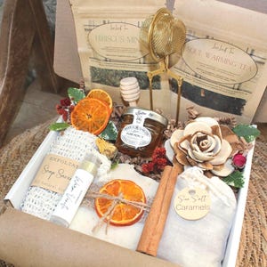 May include: A gift box containing two tea pouches labeled Hibiscus Mint and Soul Warming Tea, a gold metal tea infuser, a small jar of honey with a wooden dipper, a soap saver bag, a lip balm, a dried orange slice, a wooden flower, and sea salt caramels.