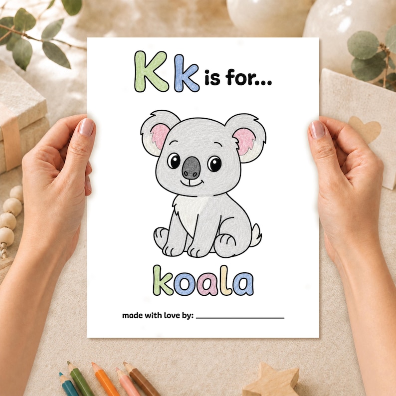 Hands holding a completed K is for koala coloring page from a printable baby shower ABC keepsake book, colored with pencils.