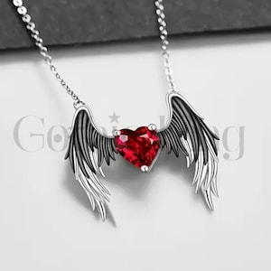 May include: A silver necklace with a heart-shaped, red gemstone pendant, flanked by wing-shaped embellishments. The chain is fine, and the pendant is the central feature. The background is a muted grey.