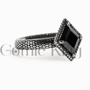 Peut inclure: Une bague en argent avec une pierre précieuse noire de forme carrée sertie dans un cadre en argent. La bande de la bague a un design texturé.