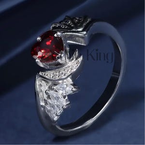 May include: A silver ring featuring a red heart-shaped gemstone and a wing design with clear cubic zirconia stones. The ring is displayed against a blue background.