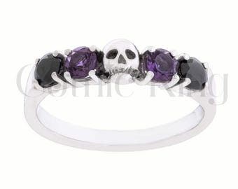 May include: A silver ring featuring a skull and four gemstones. The ring has a small skull in the center, flanked by two purple and two black gemstones. The ring is a gothic style.