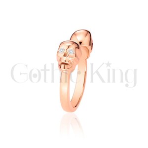 May include: Rose gold ring featuring two skull designs. The skulls have diamond-like accents in the eye sockets. The ring is a unique piece of jewelry with the text "Gothic King" in the background.