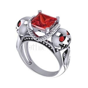 May include: A silver ring with two skull-shaped accents and a large red square-shaped gemstone. The ring has a rope-like design around the gemstone.