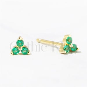 May include: Two gold-colored stud earrings, each featuring three round, vibrant green gemstones arranged in a triangular cluster. The earrings are set against a white background, highlighting their design and color.