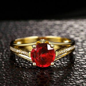 May include: A gold ring with a large, round, red gemstone as the centerpiece. The band is split, with one side featuring small, clear stones. The ring has a classic, elegant design, perfect for special occasions.