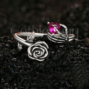 May include: A silver-colored ring with a rose and skeleton hand design. The skeleton hand holds a pink heart-shaped gemstone. The ring has a gothic theme.