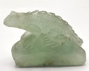 May include: A green glass sculpture of a lizard perched on a rock. The sculpture is detailed with scales and textured markings.