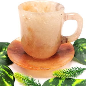 May include: A set of two, light orange, hand-carved, stone teacups and saucers. The cup and saucer are made of a smooth, polished stone. The cup has a rounded handle. The saucer is slightly larger than the cup and has a slightly raised edge.