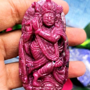 Könnte beinhalten: Ein geschnitzter Rubinanhänger, der eine Gottheit darstellt. Der Anhänger hat eine tiefrosa Farbe mit aufwendigen Details, darunter eine Krone und fließende Gewänder. Die Figur ist vor einem ovalen Hintergrund platziert.