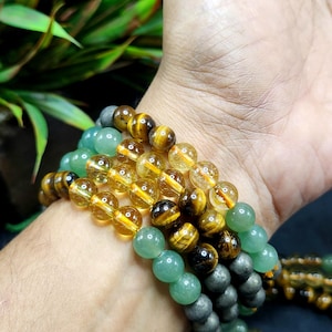 May include: A close-up of several beaded bracelets in shades of green, yellow, and brown. The bracelets are made of polished gemstones, including jade, tiger's eye, and citrine. The bracelets are stacked on a wrist.