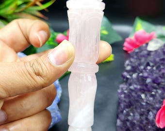 May include: A carved rose quartz crystal wand. The wand is light pink with a detailed floral design at the top and a pointed tip. The wand is held against a dark background with greenery and pink flowers.