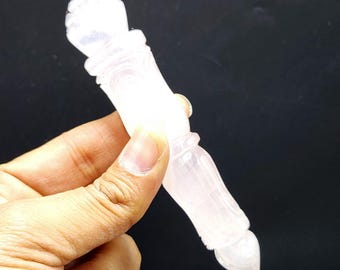 May include: A carved, light pink crystal wand with intricate details. The wand has a rounded top and a pointed bottom. The crystal is held against a black background.
