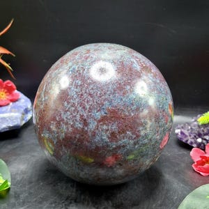 Puede incluir: Una gran esfera de piedra preciosa pulida con un patrón moteado de borgoña, azul y gris. La esfera es un punto focal, rodeada de elementos decorativos, incluyendo flores y otros cristales. La superficie de la esfera refleja la luz.