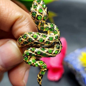 以下が含まれることがあります： 緑色のヘビのデザインが施された金の指輪。ヘビは小さな緑色の宝石でできており、金のバンドに巻き付けられています。
