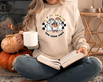 Puede incluir: Una mujer lleva una sudadera beige con un gráfico de fantasmas con sombreros y el texto "The Phantom Five Boo Crew Vibes". Está sentada en el suelo con calabazas y sosteniendo una taza blanca.
