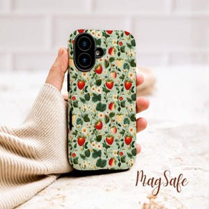 Puede incluir: Funda de teléfono con un diseño de fresas. La funda tiene un fondo verde claro con fresas rojas, flores blancas y hojas verdes. Tiene un recorte para una cámara de doble lente y el texto "MagSafe".