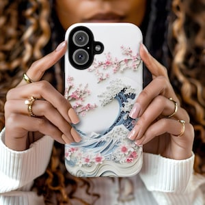 May include: White phone case featuring a Japanese wave design with cherry blossoms. The case has a 3D effect, with pink flowers and a blue and white wave pattern. The phone case has a dual-lens camera.