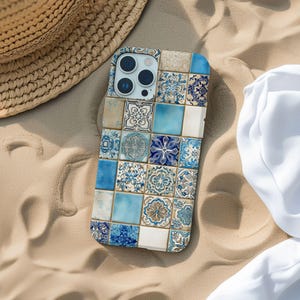 Pode incluir: Uma capa de smartphone com um design de mosaico em tons de azul, branco e bege. A capa apresenta um padrão de azulejos quadrados com motivos florais e geométricos. A capa está sobre uma superfície arenosa, ao lado de um chapéu de palha e tecido branco.