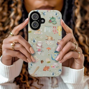 May include: A phone case with a whimsical design featuring teacups, bunnies, and other charming details. The case has a light green background with a variety of pastel colours.