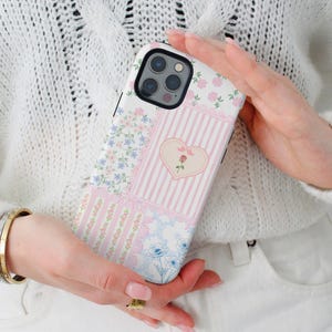 Puede incluir: Una funda de teléfono blanca con un diseño de patchwork que presenta patrones florales rosas, azules y blancos, un corazón con una rosa y un patrón a rayas. La funda está diseñada para un iPhone.