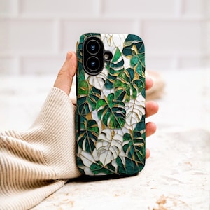 Puede incluir: Funda de teléfono con un diseño de hojas de monstera verdes y blancas con detalles dorados. La funda tiene un acabado brillante y una abertura para cámara de doble lente. El diseño tiene un tema botánico.