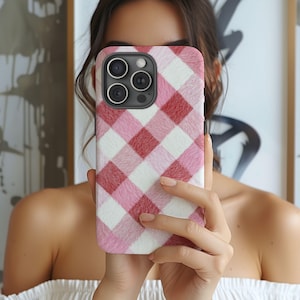 Può includere: Una custodia per telefono con un motivo a quadri rosa e rossi. La custodia ha un aspetto strutturato e peloso e un modulo fotocamera con tre obiettivi. La custodia è tenuta davanti a una persona.