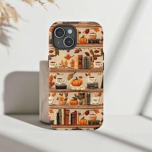 May include: A phone case with a fall-themed design featuring a bookshelf with books, pumpkins, and teacups. The background is a light brown color.