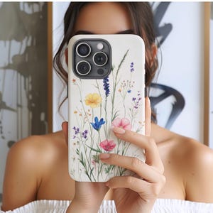 Pode incluir: Uma capa de telefone branca com um design floral em aquarela com flores amarelas, azuis e rosas. A capa tem um acabamento brilhante e um recorte para a câmera. A capa do telefone é segurada em frente a uma pessoa.