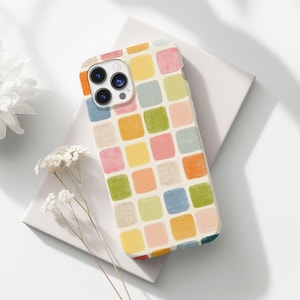 Pode incluir: Capa de telefone colorida com um padrão de grade de quadrados pastel em tons de rosa, amarelo, verde, laranja e azul. A capa está em uma superfície branca, com uma flor branca e flores secas no fundo.