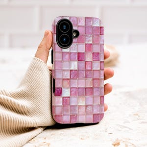 May include: A pink mosaic tile phone case with a dual-lens camera. The case features a pattern of small, square tiles in varying shades of pink, creating a textured appearance. The phone case is held in a person's hand.