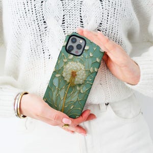 Op de afbeelding: Een teal telefoonhoesje met een gedetailleerd paardenbloemontwerp. De paardenbloem is wit en goudkleurig, tegen een achtergrond van delicate stelen en bladeren. De hoes heeft een zwarte rand.