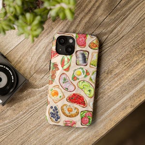 Op de afbeelding: Een telefoonhoesje met een kleurrijk patroon van verschillende soorten toast met verschillende toppings, waaronder avocado, eieren en fruit. De hoes is ontworpen voor een telefoon met een drievoudige camera lens.