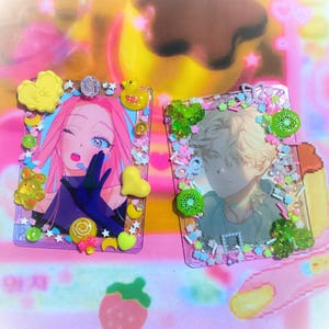 May include: Two rectangular, decorated photo cards. One features a pink-haired anime character with blue eyes, surrounded by yellow hearts, a duck, and other embellishments. The other card has a male anime character with green kiwi and other decorations.