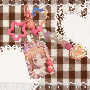 May include: A keychain featuring a rectangular card holder with an anime-style character illustration, decorated with pink star and butterfly charms, a pink bell, and a beaded strand of white, purple, and pink beads ending in a heart charm.