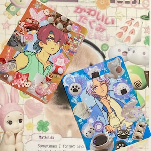 May include: Two vibrant art cards with anime-style illustrations, adorned with decorative stickers, charms, and glitter. One card depicts a red-haired character, the other a purple-haired character. The background includes a bunny figurine and Japanese text.