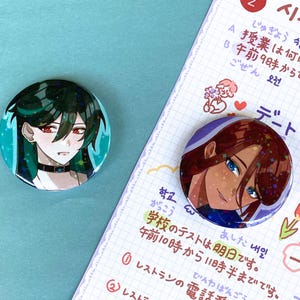 May include: Two round button pins with anime characters. The first pin features a green-haired character with red eyes and a black choker. The second pin features a brown-haired character with blue eyes. Both pins have a sparkly, iridescent background.