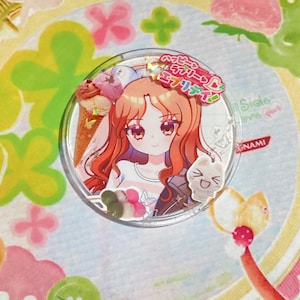 May include: A clear, circular acrylic charm featuring a cartoon girl with orange hair, holding ice cream. The charm includes Japanese text and a small cat-shaped charm. The background is a colorful, floral design.