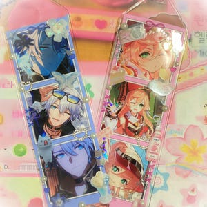 May include: Two clear acrylic bookmarks featuring anime-style character art. One bookmark has blue accents and the other has pink accents. Both are decorated with pearls and small charms. The bookmarks are on a pink and white background.