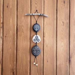 May include: A decorative wall hanging with three engraved stone pendants suspended from a piece of driftwood. The pendants are black and white, with different symbols. The background is a wooden wall.