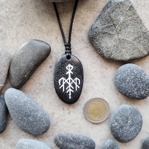 May include: A black oval pendant with a white graphic design, suspended from a black cord. The pendant is surrounded by gray stones and a coin. The design on the pendant appears to be a stylized symbol.